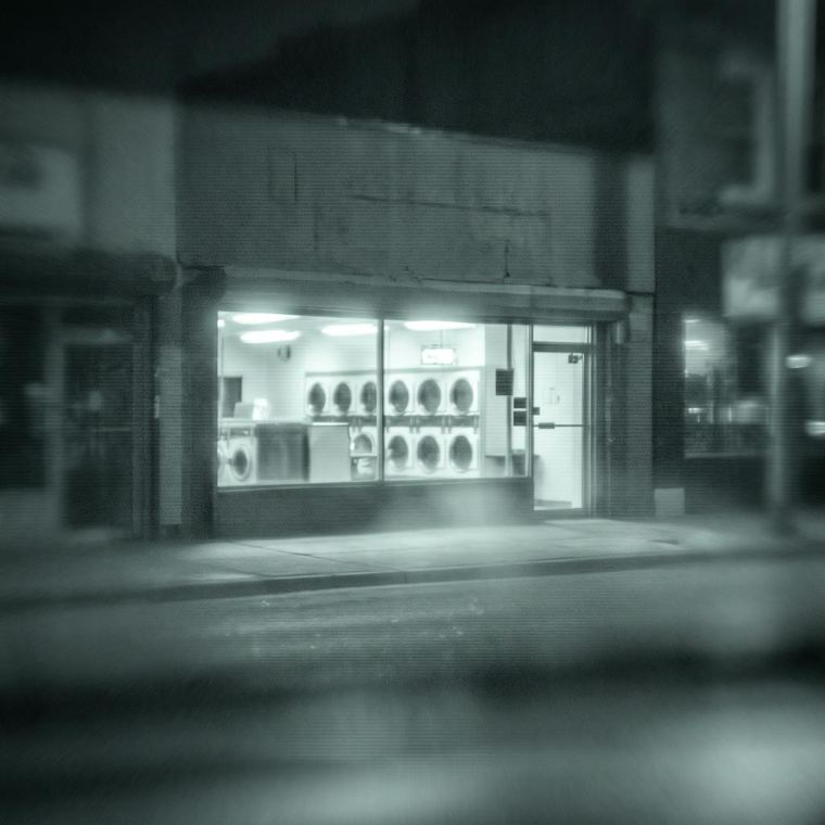 Long-lens monochrome image of a lit storefront at night, seen across a street