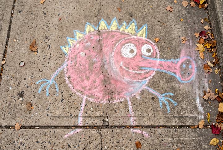 A child's chalk drawing of a large round creature with a long snout and a crown of spines, on a sidewalk scattered with leaves
