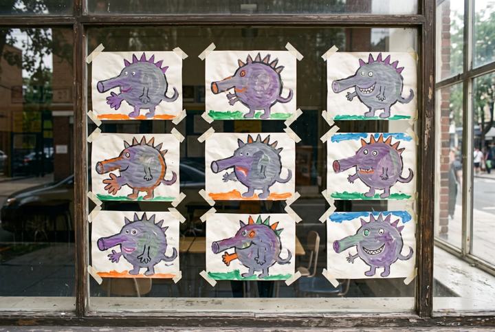 Nine children's paintings of a purple spined creature taped inside a classroom window, seen from the street