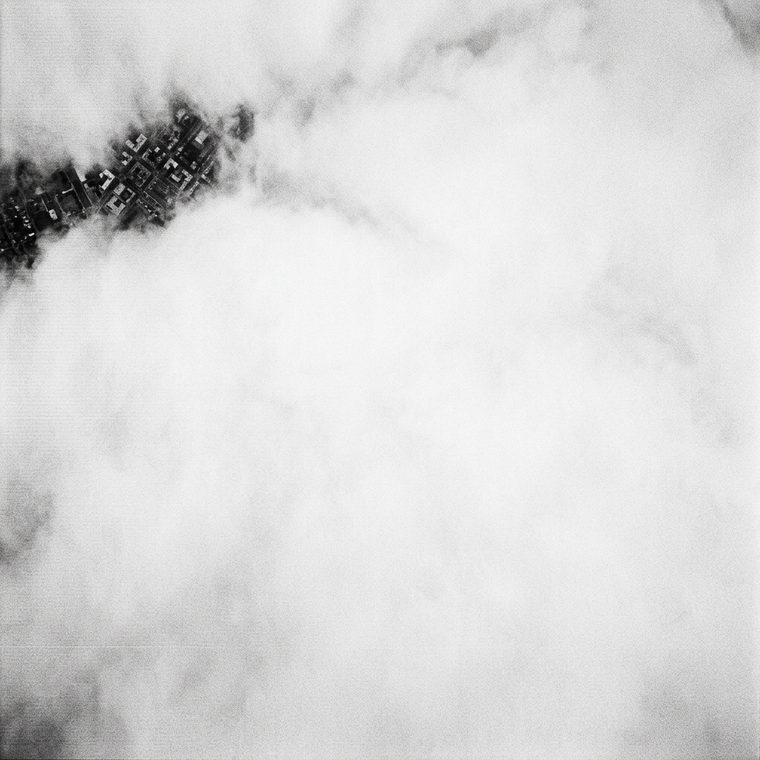 Monochrome overhead surveillance frame almost entirely filled by unbroken cloud, a small tear at one corner showing a fragment of street grid
