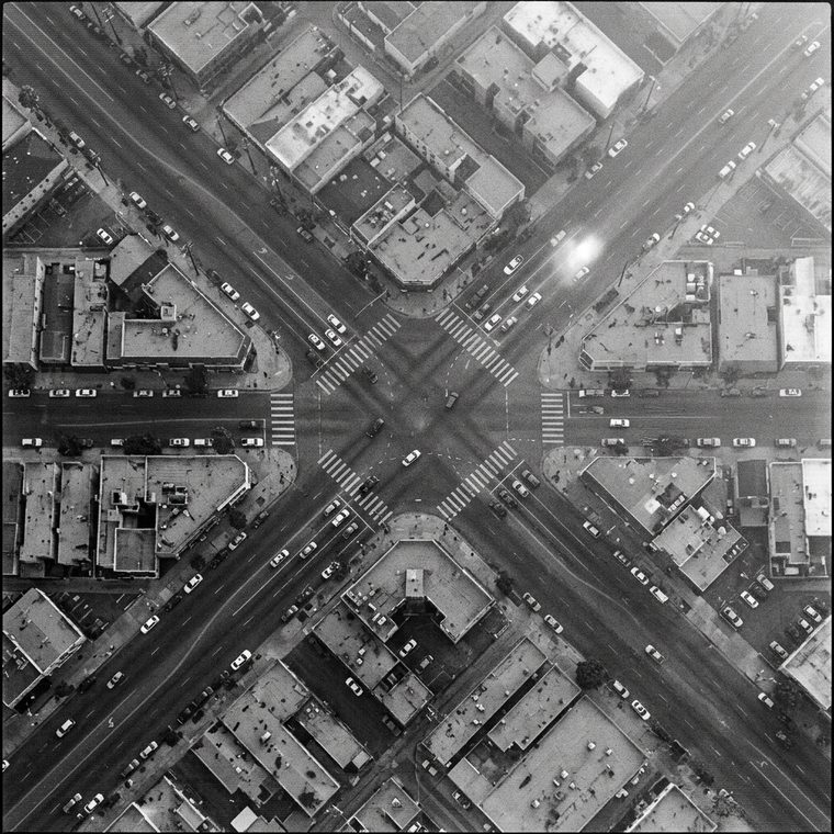 Monochrome overhead surveillance frame of a four-way city intersection, the ladder stripes of four crosswalks clearly visible