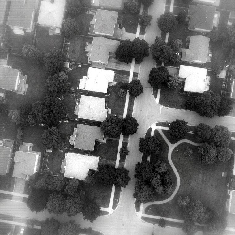 Monochrome overhead surveillance frame of a suburban block, tree canopies and a small park at one corner