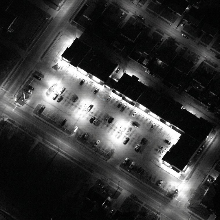 Monochrome overhead night surveillance frame of a commercial strip, pooled lamplight over a parking lot